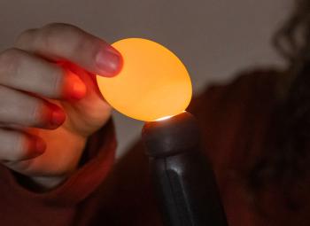 A person holds an egg over a small light source, candling it to check the interior by shining light through the shell.