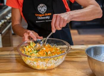A person wearing an Oregon State University Extension Service apron mixes shredded carrots, zucchini and corn in a glass bowl on a wooden counter.