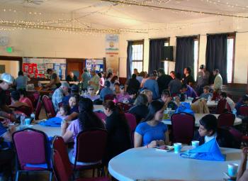 Community members gather around round tables inside a meeting hall, sharing a meal and information during a wildfire preparedness event.