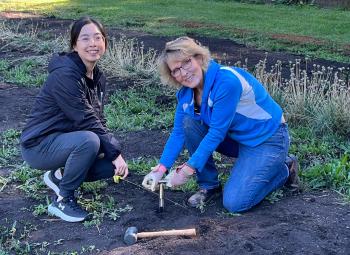 Two women kneel in a garden plot, using a soil probe to collect a sample, with a small mallet resting on the ground nearby.