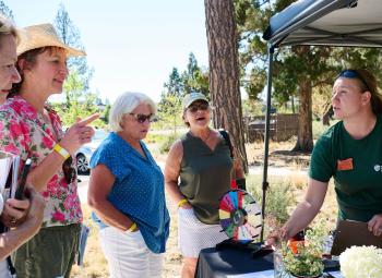OSU Extension Master Gardener volunteer talks with a small group of adults at an outdoor booth during the 2025 High Desert Garden Tour, using a display and materials to answer questions.