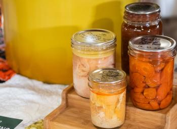 Several glass jars of home-canned foods, including carrots, sit on a wooden tray beside a large pot used for canning.