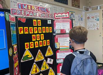 A student with a backpack looks at a classroom display board about emergency preparedness that reads “Be prepared, not scared,” with hazard symbols and information panels.