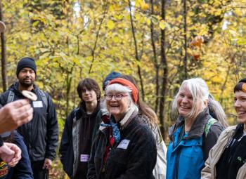 A group of adults listen and smile as a man holds up a mushroom during a guided walk in a wooded area with fall foliage.