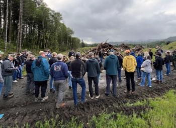 Group of adults stands on a muddy logging road listening to a speaker during a forestry field tour, with piles of cut wood and a forested hillside in the background.