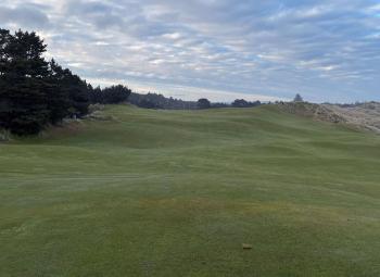 Wide view of a gently rolling golf course bordered by trees and sandy dunes under a cloudy sky.
