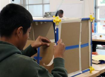 A youth works at a classroom table assembling a cardboard model structure during a hands-on career exploration activity.