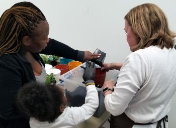n adult and child pour potting soil into containers while another instructor helps during a hands-on Seed to Supper gardening activity indoors.