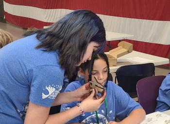 A teen mentor helps a child assemble a small wooden project during Military Kids Club camp, with other youths working nearby in front of a large American flag backdrop.