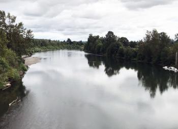 A calm river reflecting the cloudy sky is bordered by dense green trees on both sides, with a small dock visible to the right.