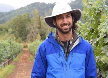 A man in a blue jacket and wide-brimmed hat stands smiling in a vineyard, surrounded by rows of grapevines and backed by forested hills.