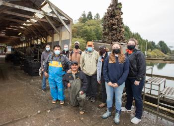 A group of people wearing masks pose for a photo in front of a net full of harvested oysters at Oregon Oyster Farms in Newport, Oregon.