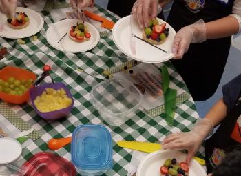 People wearing gloves assemble fruit pizzas on paper plates at a table covered with a green checkered tablecloth and containers of assorted fruit.