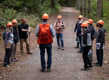 People are standing in a semi-circle on a forest trail listening to a tour guide. They're wearing orange plastic hard hats.
