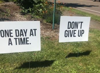 Two white yard signs with bold black lettering read “ONE DAY AT A TIME.” and “DON’T GIVE UP” on a grassy area beside a flower bed and sidewalk.