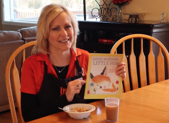 A woman sitting at a dining table holds up a children's book titled *Good Morning, Little Fox* while smiling at the camera, with a bowl of cereal and a glass of milk in front of her.