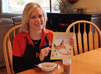 A woman sits at a kitchen table at breakfast and holds up a book that she will read to children in a video.