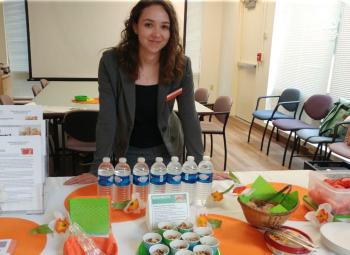 A young woman is standing behind a table that has bottled water and fresh fruit and vegetables.