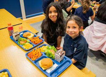 Two young girls smile at a cafeteria table with trays of colorful school lunches including burgers, sweet potato fries, fruit, salad and milk.