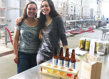 Two young women pose in front of bottles of kombucha in a brewery.
