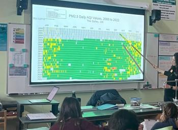 Lauren Kraemer points to a colorful air quality chart projected on a screen in a classroom, while middle school students watch attentively.