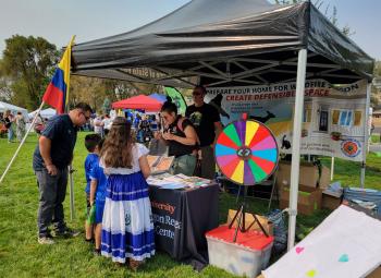 Sure! Here's a one-sentence alternative text description for the image:  "People gather at a community fair booth with wildfire preparedness materials, a colorful prize wheel, and a Colombian flag, while a woman in traditional dress talks to exhibitors."