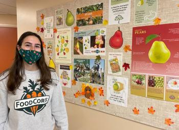 A young woman wearing a facemask stands next to a bulletin board in an elementary school hallway. She's wearing a sweatshirt that says FoodCorps on the front with a picture of carrots..