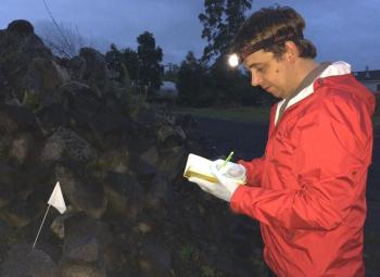 A researcher wearing a red jacket, gloves, and a headlamp takes notes in a field at dusk next to a rock pile marked with a small white flag.