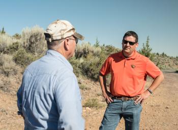 Two men have a conversation outdoors on a dirt path in a dry, shrub-filled landscape under a clear blue sky.