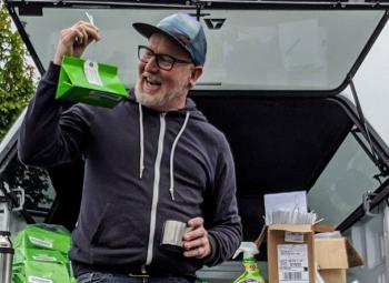 A smiling man sits on the tailgate of a truck, enthusiastically holding up a green insect trap with boxes of supplies around him.