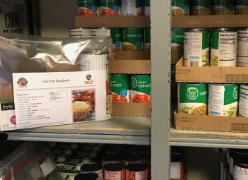Shelves stocked with canned vegetables surround a plastic bag meal kit labeled “One Pot Spaghetti” that includes ingredients and a printed recipe.
