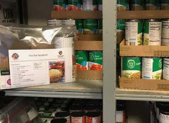 A meal kit sits on a shelf among canned food in a food pantry.